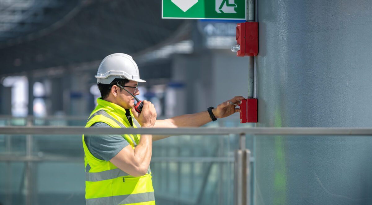 Inżynier w kombinezonie ochronnym i kasku sprawdza system alarmowy przeciwpożarowy w zakładzie przemysłowym.