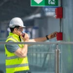 Inżynier w kombinezonie ochronnym i kasku sprawdza system alarmowy przeciwpożarowy w zakładzie przemysłowym.